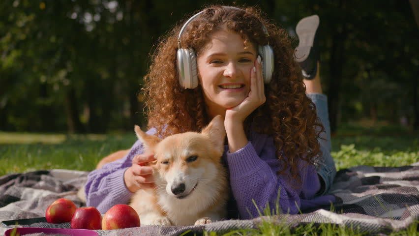 Caucasian smiling happy girl curly hair listen music in headphones relaxing summer picnic nature young pet owner handler woman rest outdoors with cute puppy welsh corgi dog best friends in park city Royalty-Free Stock Footage #3440218881