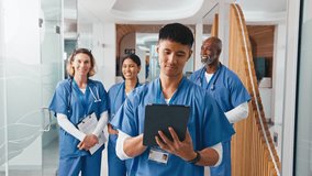 Close up portrait of smiling male doctor or nurse with digital tablet wearing scrubs standing in hospital corridor with multi-cultural team of colleagues in background - shot in real time - Powered by Shutterstock - Get 15% off with code: PIKWIZARD15