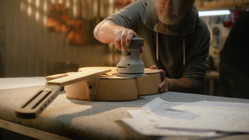 Mature Caucasian man in protective mask grinds guitar body using grinding machine. Professional carpenter makes guitar. Craftsman work in stylish workshop. Handmade and entrepreneurship concept.