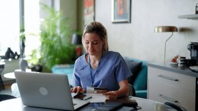 Nurse paying bills online, sitting in living room, working on notebook. Doctor in scrubs dealing with work issues from home, after workday, telemedicine consultation. - Powered by Shutterstock - Get 15% off with code: PIKWIZARD15