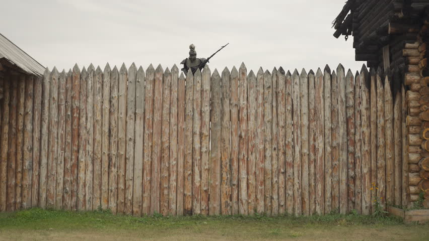 Man in armor holds spear and walks on rain on platform stretching along high log picket fence waiting for attack of enemy army. Birds fly into cloudy sky ominously