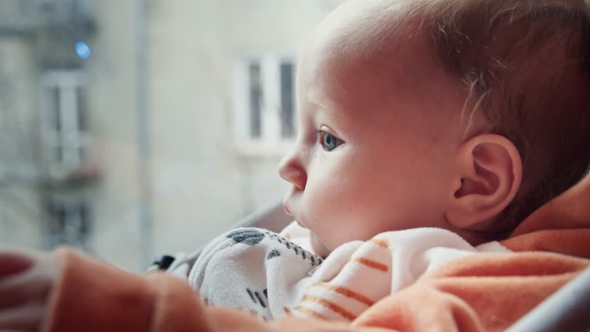 A serene baby contemplates the view from a window, bathed in the soft glow of natural light. The baby's profile is gently outlined against the blurred backdrop of urban architecture. Royalty-Free Stock Footage #3440674195
