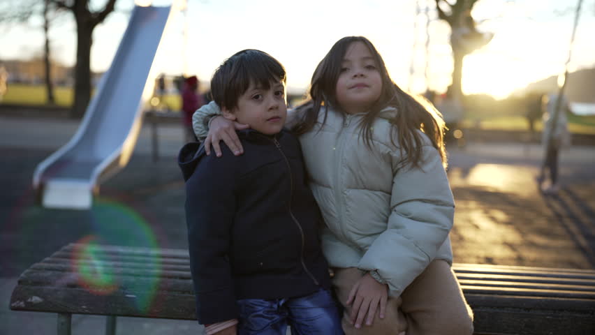 Little brother and sister seated at park bench during sunset time, backlight with flare. 5 yer old boy and 8 year old girl wearing coats, blurred children in background playing Royalty-Free Stock Footage #3440796121
