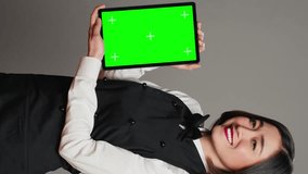 Vertical Video Woman waitress holding tablet with greenscreen display on camera, presenting blank mockup template in studio. Asian restaurant employee showing device running chromakey screen. Camera A
