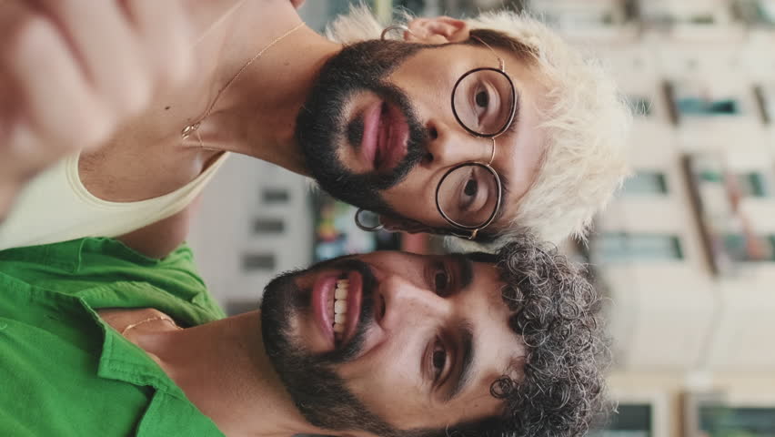 Vertical video, Gay couple stands hugging and makes video call from mobile phone, waving in greeting