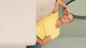 Vertical video, Bald woman dressed in yellow t-shirt and jeans sitting in living room, enjoying free time on weekend