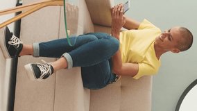 Vertical video, Smiling bald woman resting on sofa in living room writing SMS with her mobile phone