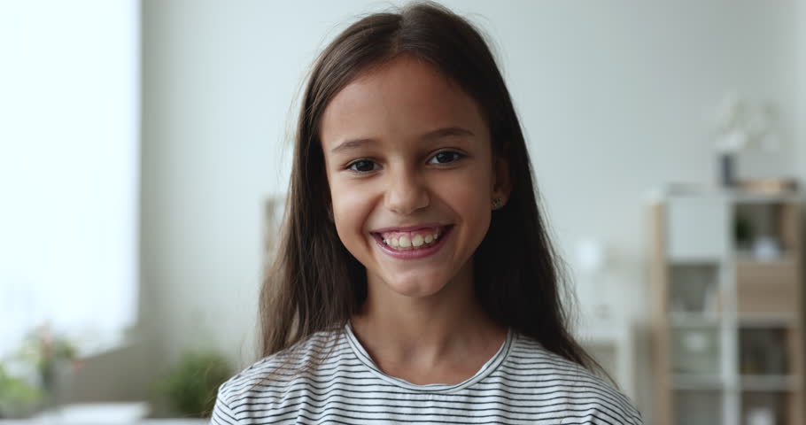 Close up head shot of cheerful beautiful little pre-teen 8s-10s girl standing in living room smile look pose for camera at home. Portrait of happy young Z or Alpha generation kid, carefree childhood