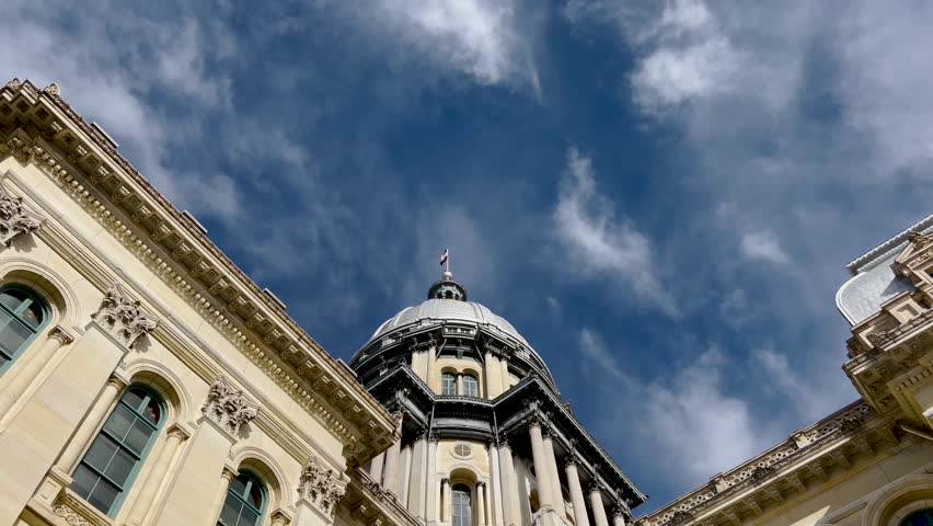 Rear views of the Illinois State Capitol Building in Springfield, Illinois, USA. Cloudy blue skies overhead. The US flag, State of Illinois Flag, and Black History Month Flag fly atop the dome.