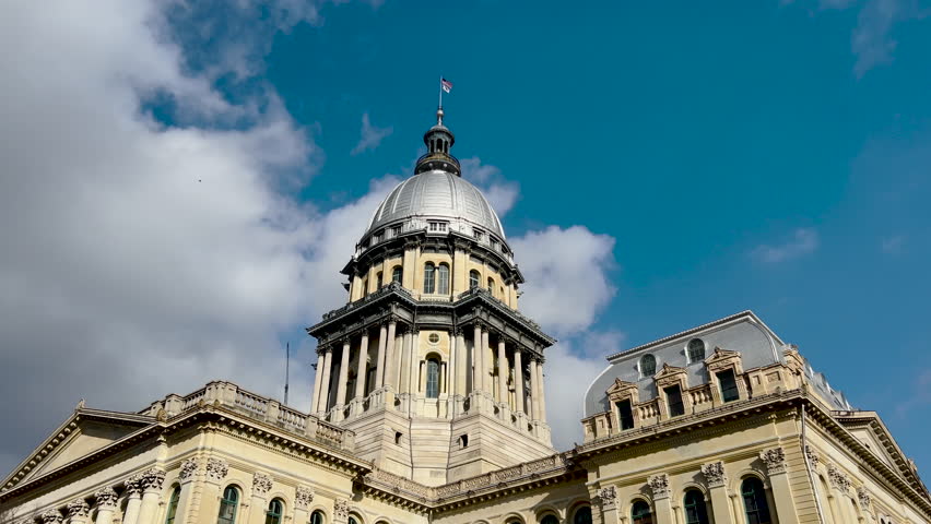 Rear views of the Illinois State Capitol Building in Springfield, Illinois, USA. Cloudy blue skies overhead. The US flag and State of Illinois Flag fly. Construction crane in the background of view.