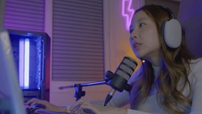 Happy Asian streamer female talking with microphone in gaming room, young gamer woman wear headphones playing video games online on computer she live stream and chat with fans, game streaming concept