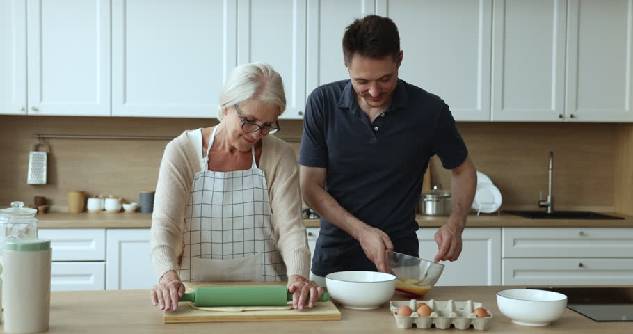 Millennial man whisks eggs for pastries talk to mature positive mother flattening dough for homemade holiday pie, cooking in the kitchen. Time together with older relative, understanding, family bond Royalty-Free Stock Footage #3440926601