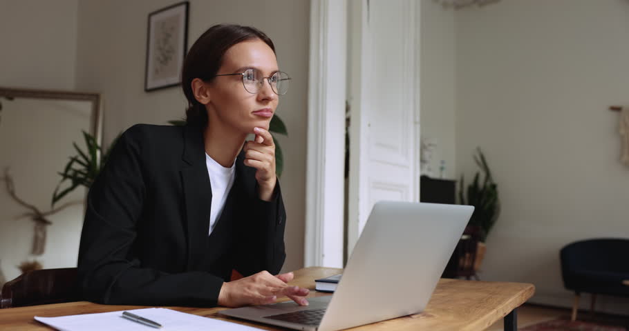 Positive young business professional woman in glasses thinking at workplace, typing on laptop, browsing Internet, smiling, touching chin, working on startup project presentation computer Royalty-Free Stock Footage #3440944013
