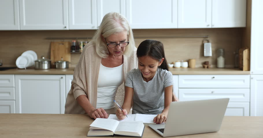 Loving retired 60s granny explaining homework to pretty little 10s grandchild. Cute school age girl doing assignment with help of grandmother in kitchen at home. Education, family support, tutoring Royalty-Free Stock Footage #3440951797