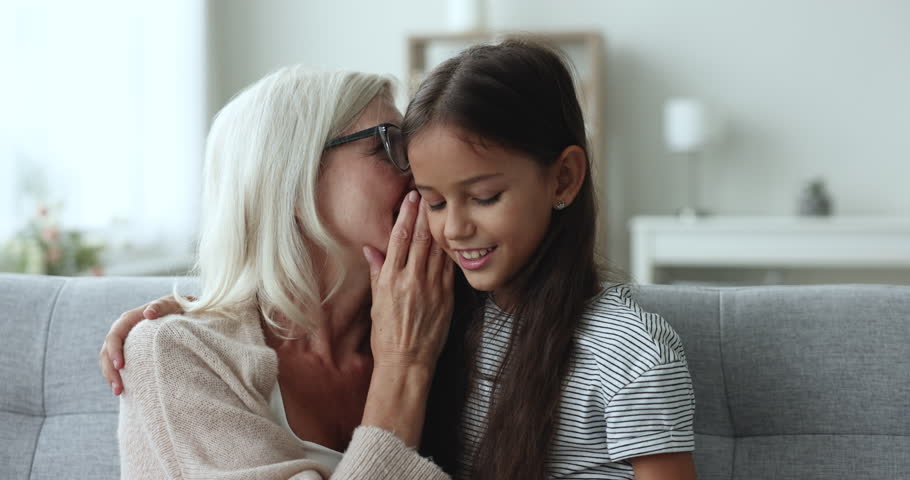 Pretty 10s granddaughter spend weekend time with old attractive granny sitting on couch, girl whisper girlish secrets in ears, secretly talking to grandma, enjoy gossips. Family relationship, trust Royalty-Free Stock Footage #3440953251