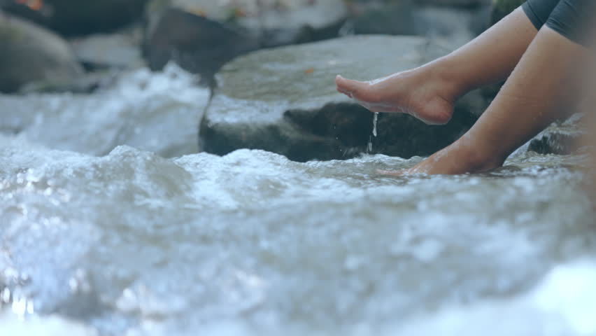 Woman sitting beside waterfall and dangling legs into water and splashing water with fun. People travel and enjoy with nature. Slow motion shot. Slow motion shot.