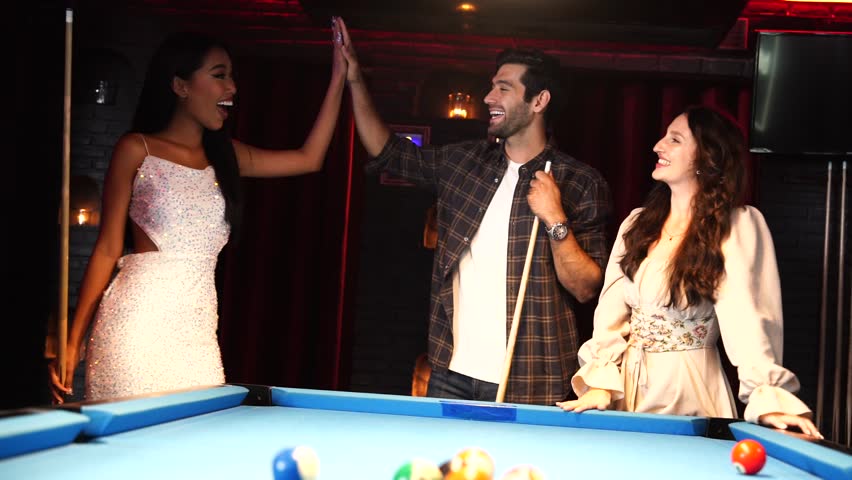 Group of multi-national friends, one man introduces playing pool at a pool table to a Caucasian woman and an Asian woman, fun and exciting, competing at a pool table in a nightclub.
​