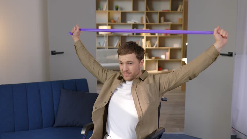 Young disabled man in wheelchair doing exercises with rubber band at home. Caucasian handicapped guy working out in living room