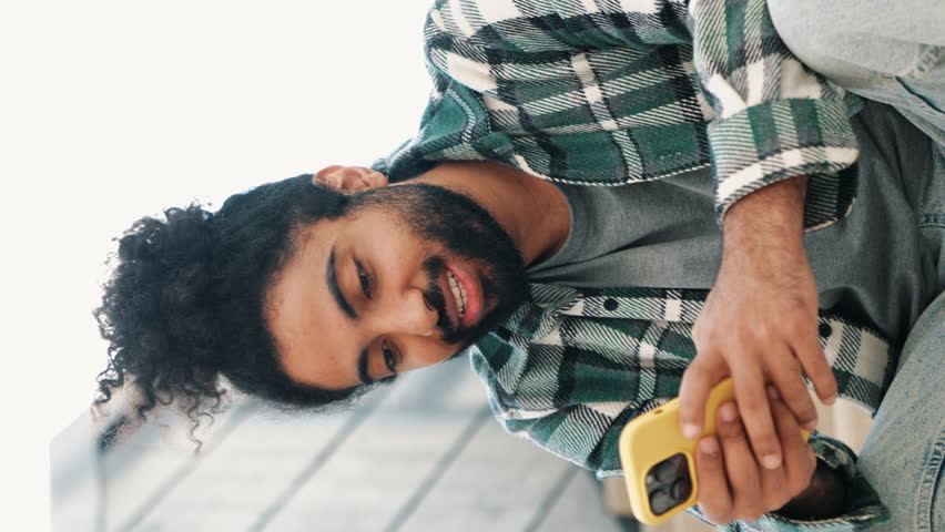 Vertical video. Handsome smiling hipster model. Unshaven Arabian man in summer casual clothes. Fashion male with long curly hairstyle posing in the street. Holds smartphone, uses phone mobile apps. Royalty-Free Stock Footage #3441200375