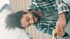 Vertical video. Handsome smiling hipster model. Unshaven Arabian man in summer casual clothes. Fashion male with long curly hairstyle posing in the street. Holds smartphone, uses phone mobile apps.