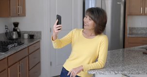 Mature woman smiling enjoying talking using video chat technology on smartphone laughing relaxing standing in home kitchen. Cheerful attractive female online using internet mobile phone leaning.

