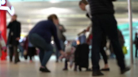 Little girl standing next to suitcase and looks on passing passenger and trolley at airport in blur. Family waiting at international airport terminal. Crowd of people at airport. Mother and daughter