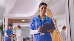 Camera tracks female doctor or nurse wearing scrubs using digital tablet in busy hospital corridor with colleagues in background - shot in slow motion - Powered by Shutterstock - Get 15% off with code: PIKWIZARD15