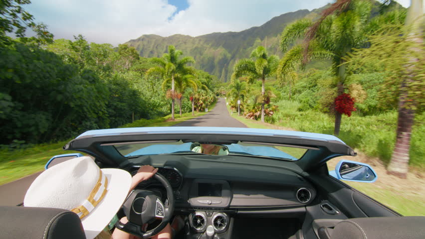 Happy girl in blue convertible car enjoying summer vacation, waiving hand out of the car and showing shaka hawaiian sign. Woman travels by tropical island with tropical exotic mountain landscape Oahu Royalty-Free Stock Footage #3441512621