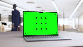 Laptop with a green chroma key screen on a wooden desk in a modern office, with a blurred person in the background.