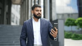 Adult bearded businessman talking on a video call using a phone standing near an office building. Smiling handsome entrepreneur having an online conversation with a friend, client or business partner