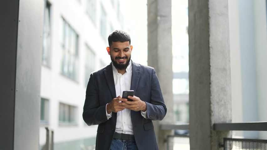 Happy smiling businessman in formal suit is using phone walking near an office building. Handsome bearded male texting, chatting online, browsing web social media, reads writes messages on smartphone