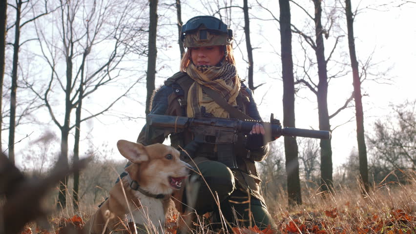 Young pretty redhead woman in military uniform armed with rifle playing with dog