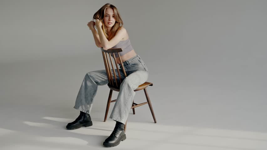 Young beautiful woman model in casual clothes sitting on chair and posing on camera at studio with white background. Attractive female in crop top and jeans