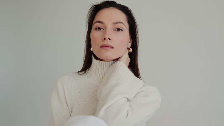 A woman in a white sweater and earrings is gently touching her cheek with a smile, showcasing a human body gesture of comfort and femininity. Face lifting, cosmetics. Portrait of beautiful young woman
