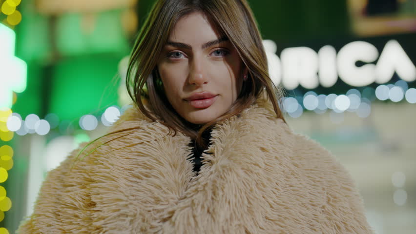 A woman with layered hair, wearing fur coat, stands in front of a store with a happy smile on her face. Her eyelashes flutter as she looks at the waterthemed event happening nearby