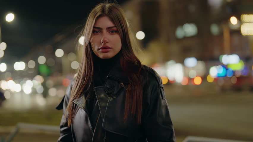 A stylish young woman stands confidently on a busy urban street illuminated by night lights, showcasing contemporary fashion.