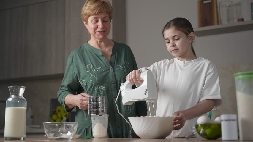 Lovely grandmother teach smiling granddaughter to make dough for pancakes for breakfast. Having fun and enjoying time together on family kitchen at home on women's day Royalty-Free Stock Footage #3441842103