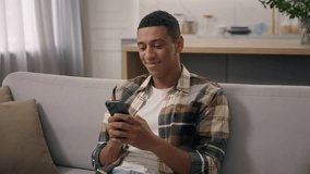 Happy smiling African American man relax on sofa at home smile laugh looking mobile phone screen smartphone chat browsing telephone ethnic guy biracial male watching funny video online fun laughing