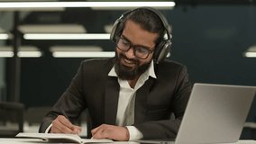 Happy smiling 30s Arabian businessman in headphones looking at laptop watching educational seminar class lesson video call in office Indian business man online negotiations writing notes in copybook