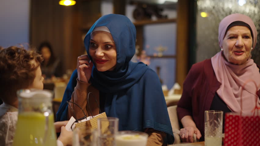 Young Muslim Mother Talking to Her Son Over the Eid or Ramadan Gift She Gave Him While Sitting at the Table in the Restaurant With Other Family Members Royalty-Free Stock Footage #3441966535