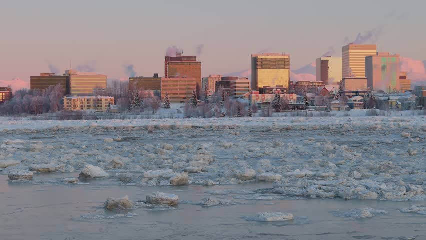 Slowly drone dolley shot over a frozen lake with in the background the skyline of Anchorage illuminated by sunset.