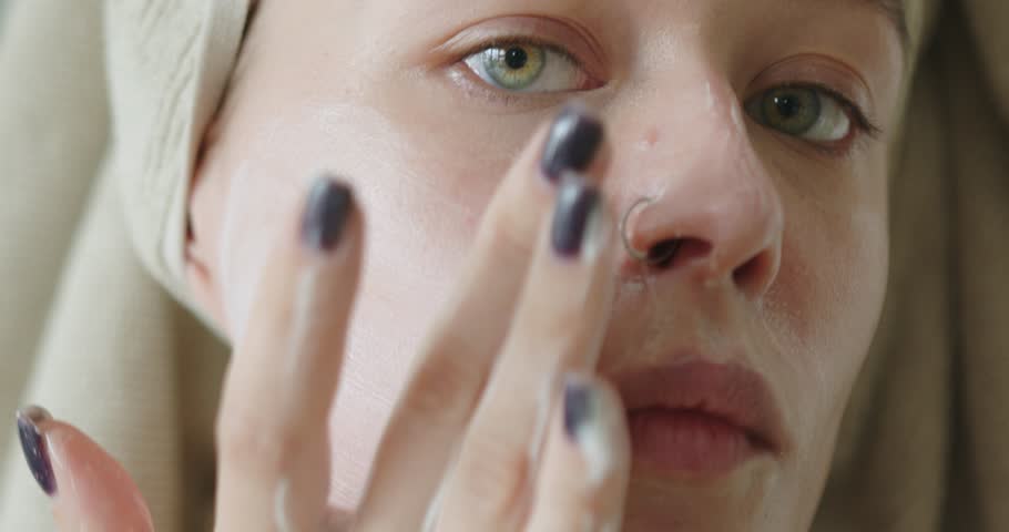 The image is a close-up of a young woman's face as she gently applies facial cream. Her green eyes are focused, and her expression is calm. The cream on her fingers and the sheen on her skin suggest Royalty-Free Stock Footage #3442104287
