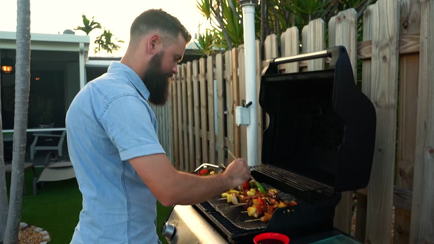 Caucasian male basting chicken, steak, vegetable shish kabob skewers on backyard grill