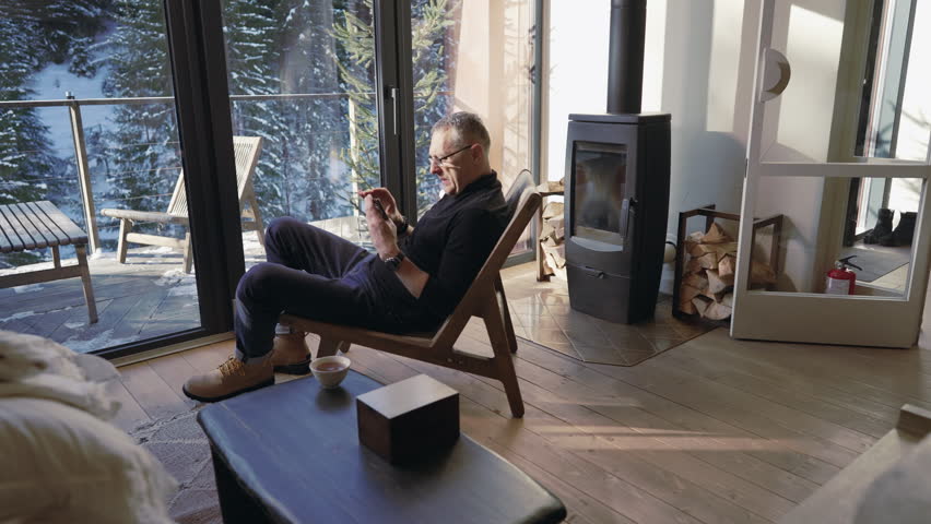 A man drinks tea while enjoying the view from the window of his country chalet.