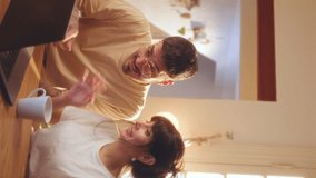 Young cheerful couple watching funny video on the Internet on laptop, laughing, kissing and hugging during the day at home. Vertical format clip