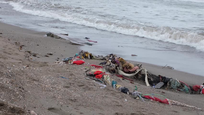 Piles of discarded clothes and fabric, fast fashion, washed ashore onto sandy beach from ocean with gentle waves on a tropical island destination