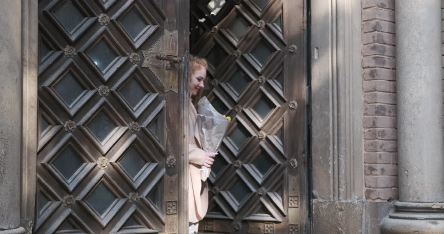 Joyful young blonde woman in beige coat walks out of building with massive wooden doors. Girl holds bouquet of flowers and smiles.