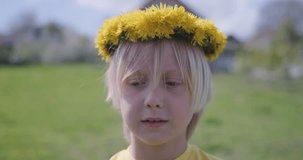 Close up video portrait of blond 10-years boy outside. Fair-haired child wears wreath of dandelions.