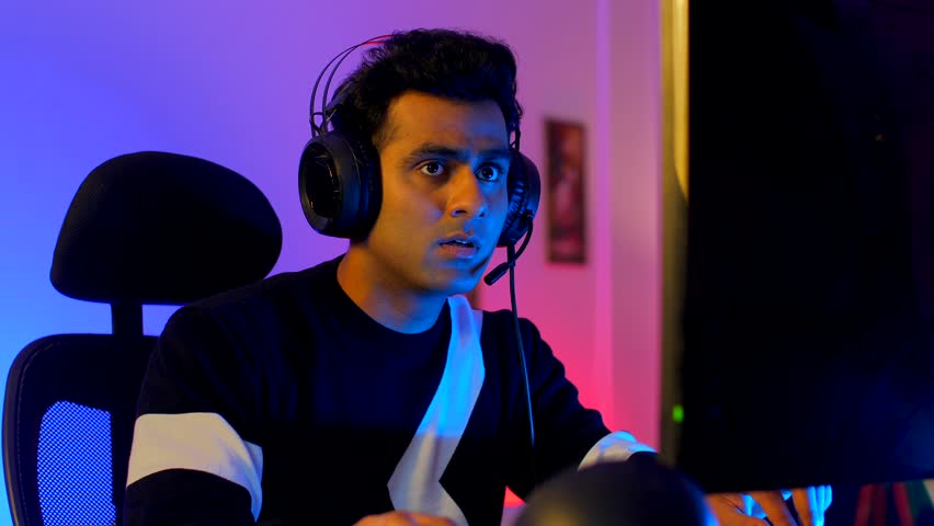 An upset young Indian man playing an esports tournament - feeling sad and disappointed after being defeated. An Asian gamer wearing headphone shows mixed emotions while using the computer - neon li... Royalty-Free Stock Footage #3442415337