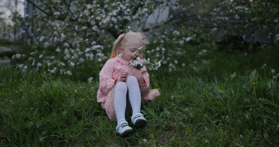Cute spring fashion girl under blossom apple tree. Fair-haired child girl in pink outfit sits in flowering garden and blowing on flower petals.